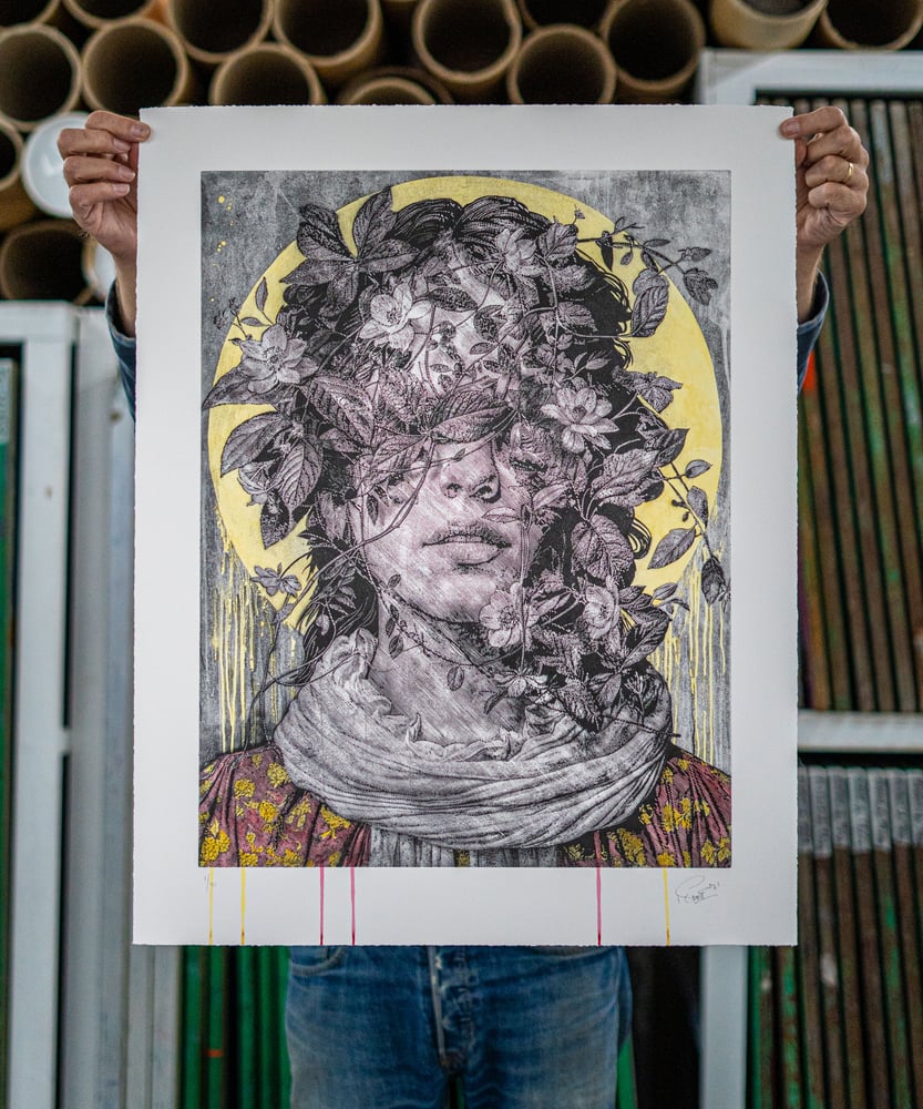 SNIK’s AFTERDAWN main-edition etching held up in the studio, showing a flower-covered portrait with yellow and pink hand-finishing.