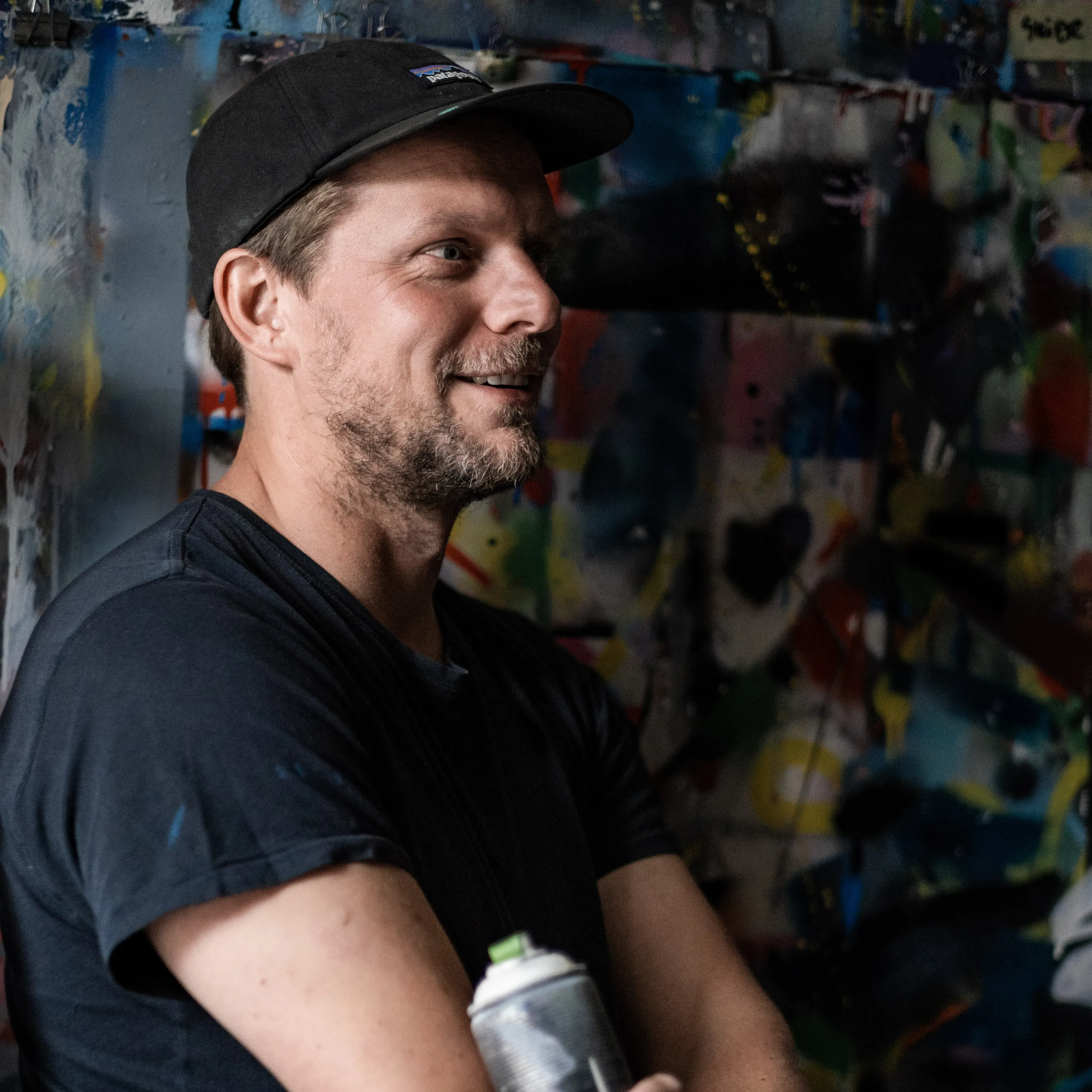 Martin Whatson in a black cap and T-shirt holding a spray can in front of a paint-covered studio wall.